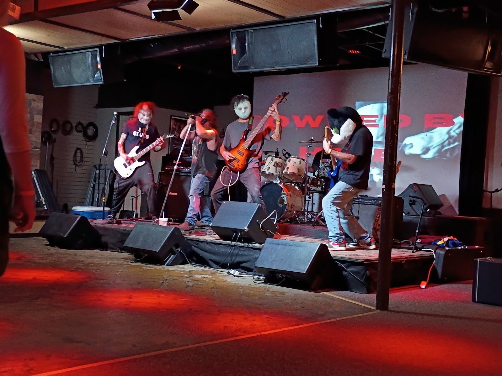 A five-piece band in Halloween masks playing on the Haltom Theater's low stage under red lights, with a projection screen and PA speakers behind them