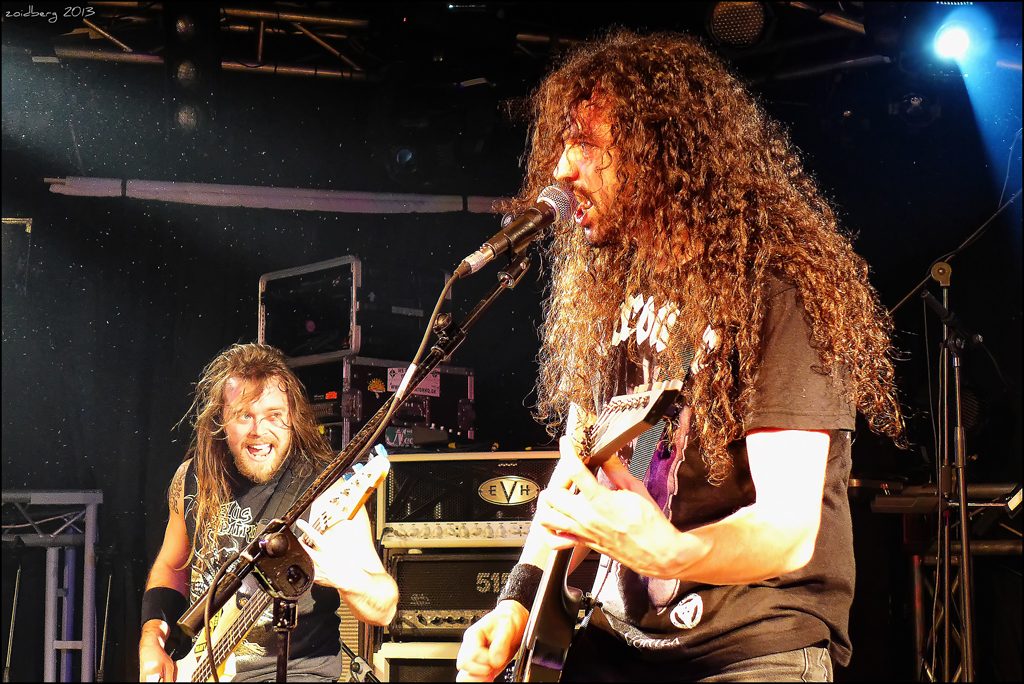 Havok's curly-haired singer and guitarist David Sanchez sings into a microphone while a grinning bandmate plays beside him on a dark club stage