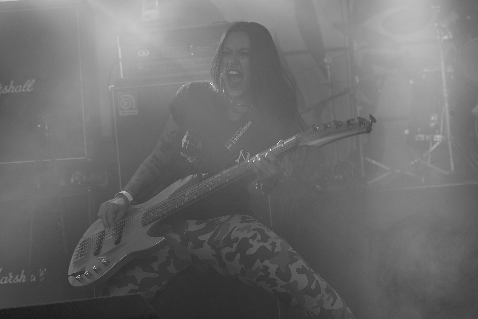 Black and white photo of Nervosa's bassist screaming mid-song through stage haze, a bass guitar angled across the frame in front of Marshall and Ampeg cabinets