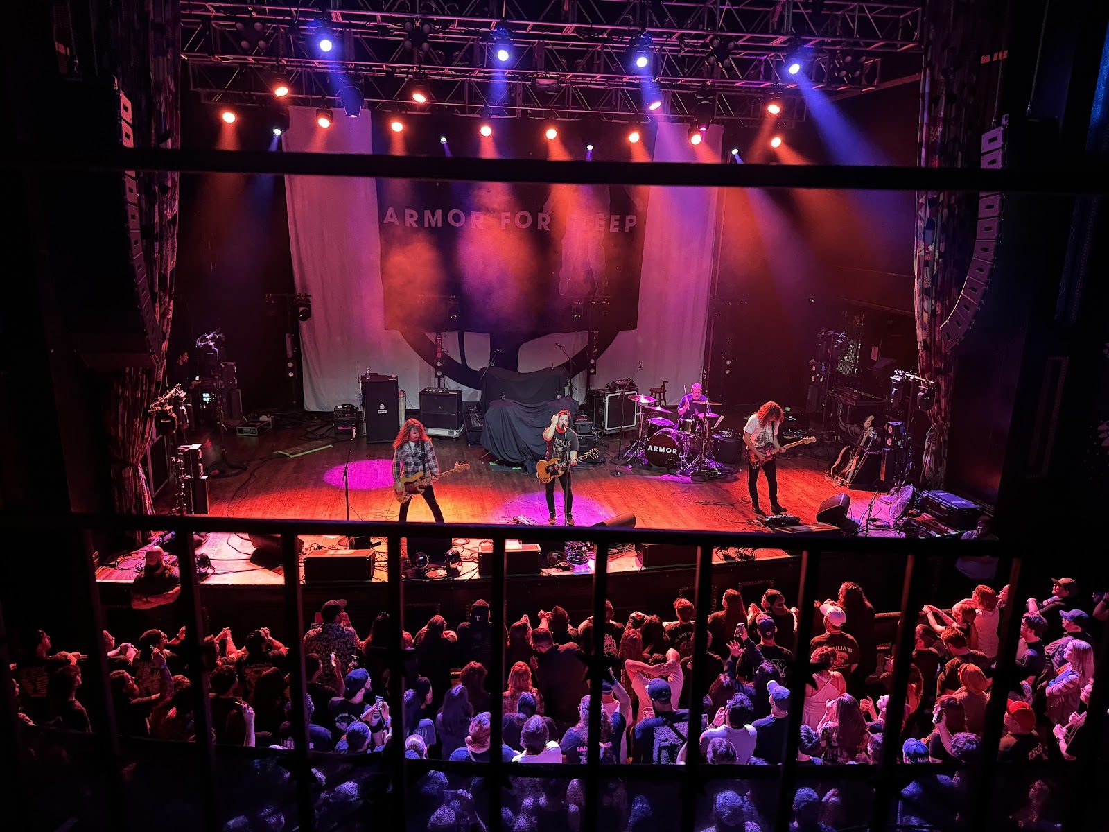 View from the House of Blues Dallas balcony through a railing to a four-piece band under orange and purple lights, a packed floor below