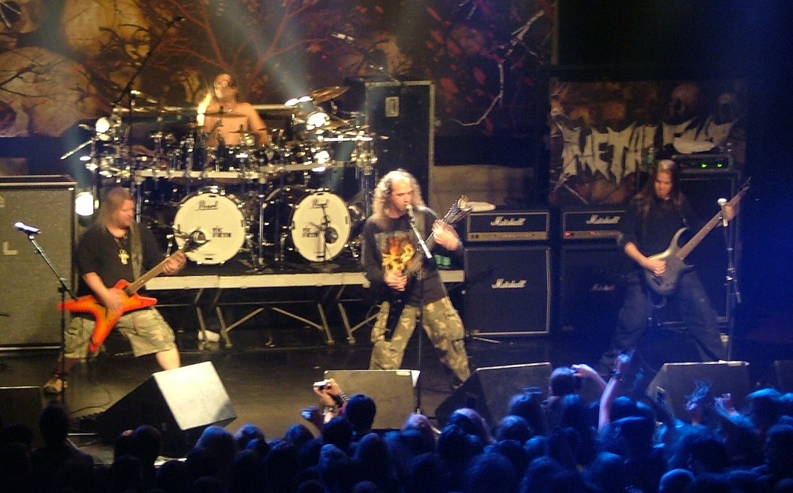 Nile on a club stage, two guitarists and a bassist in front of Marshall stacks and a large drum kit, hands raised in the crowd below