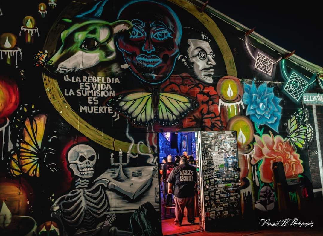 Reno's Chop Shop's open side door at night, framed by a mural of a skeleton, butterflies and a glowing face, with a man in a Reno's Security shirt standing in the doorway