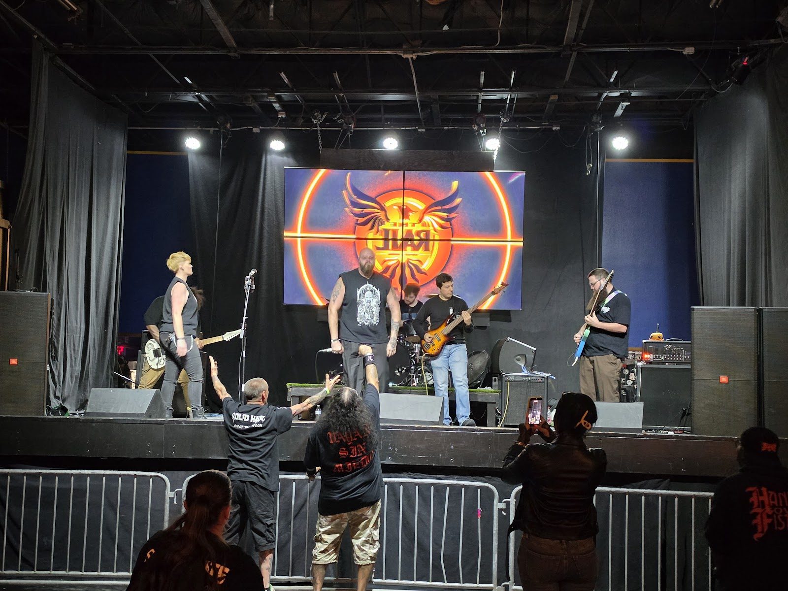 A five-piece metal band on The Rail's wide black stage under white lights, The Rail's winged logo glowing on a video wall, fans at the barricade raising their arms