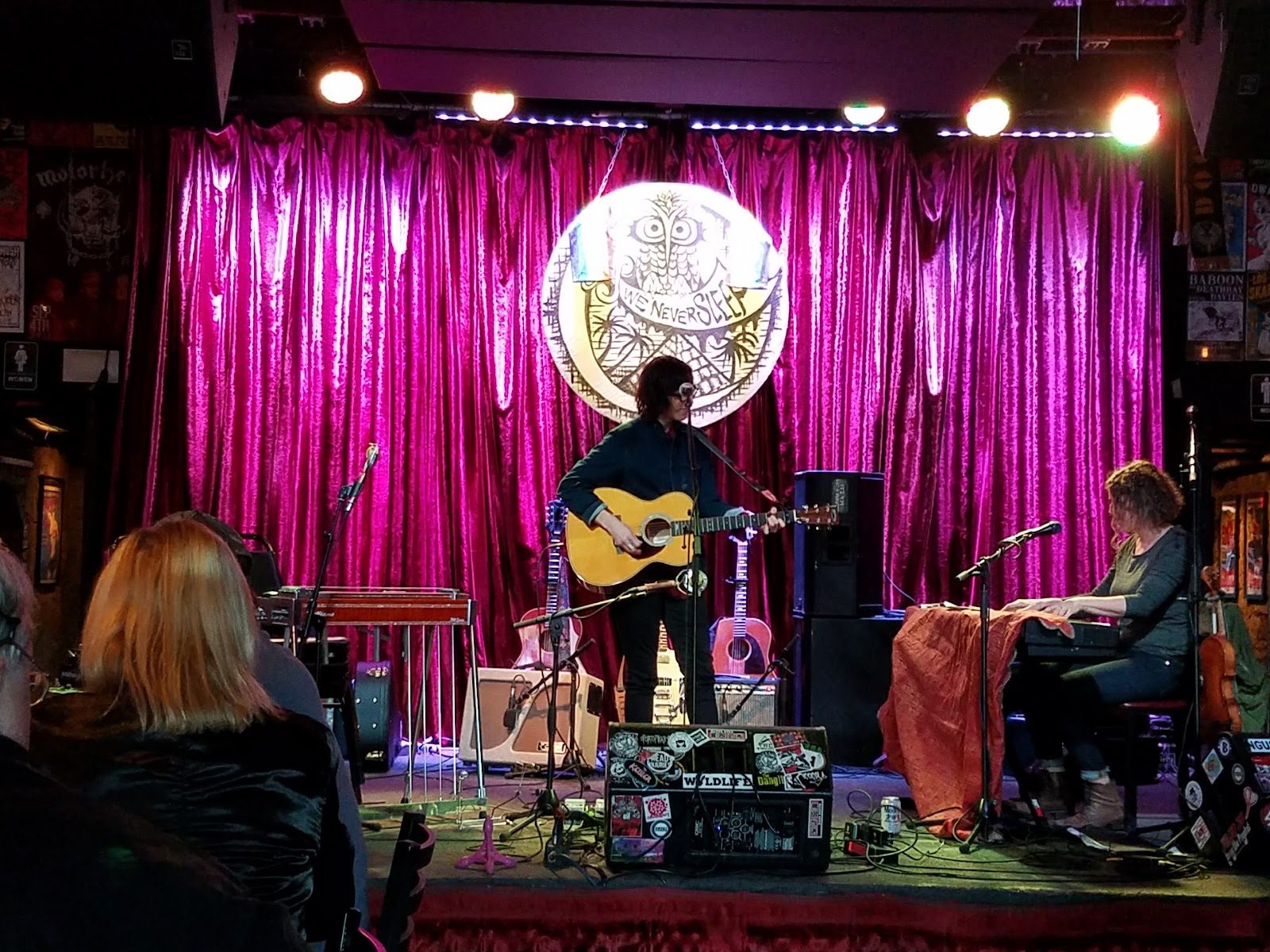 A singer with an acoustic guitar and a keyboard player on Three Links' small stage, in front of a pink velvet curtain and a round owl sign reading We Never Sleep