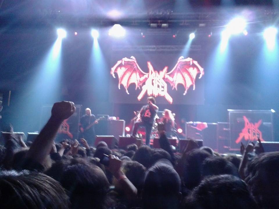 Dark Angel on a concert stage under blue spotlights, the red winged band logo glowing on a screen above a packed crowd with raised fists