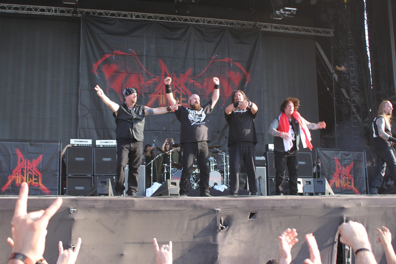 Dark Angel's members raising their arms to the crowd on a festival stage, the band's red winged logo on the backdrop and fans' devil-horn hands in the foreground