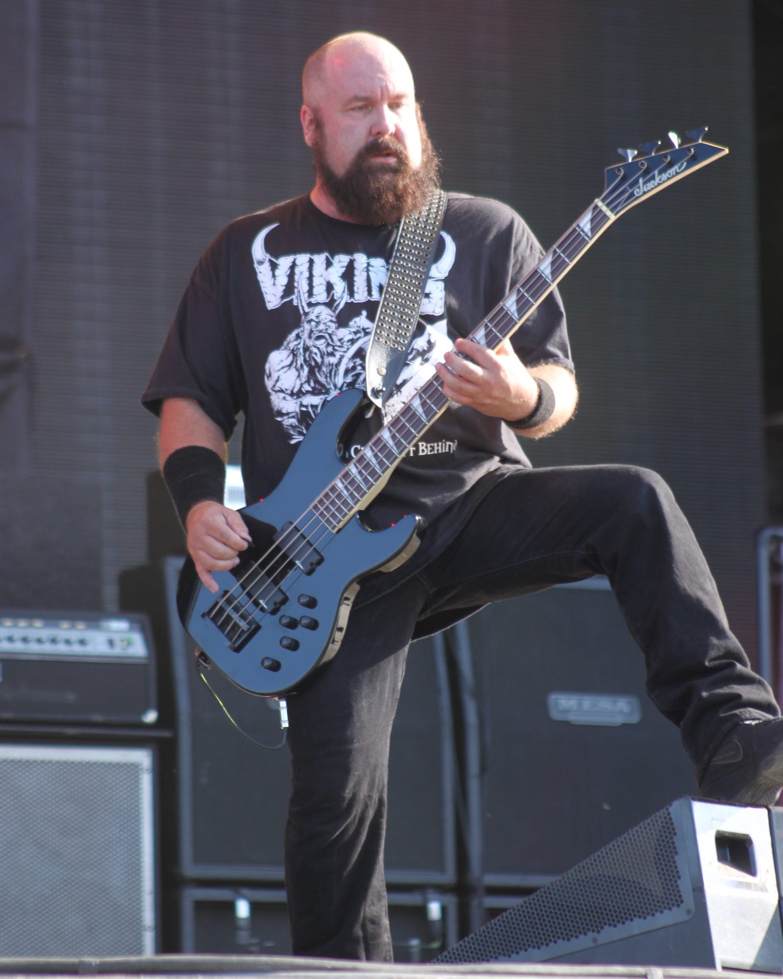 Bald, bearded bassist Mike Gonzalez in a Viking T-shirt with one foot on a monitor, playing a blue bass on a festival stage