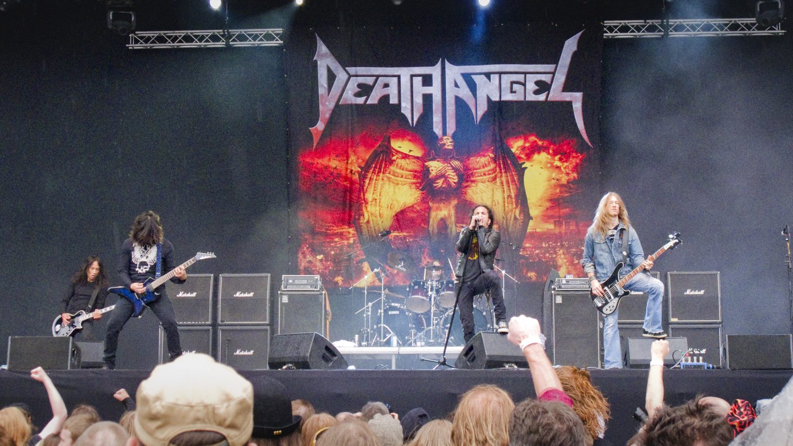 Death Angel playing an outdoor festival stage under a huge Death Angel logo backdrop: two guitarists at left, the singer at a center microphone, a bassist at right and the drummer behind, with fans' raised fists in the foreground
