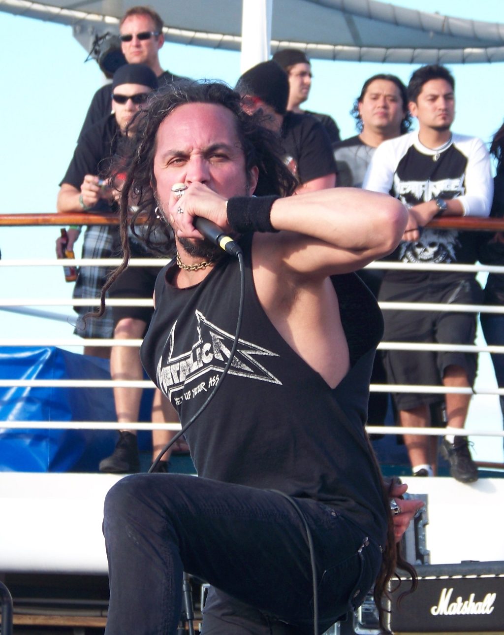 Mark Osegueda, with long dreadlocked hair and a sleeveless Metallica shirt, crouching and singing into a microphone on a ship's deck while fans watch from the rail behind him