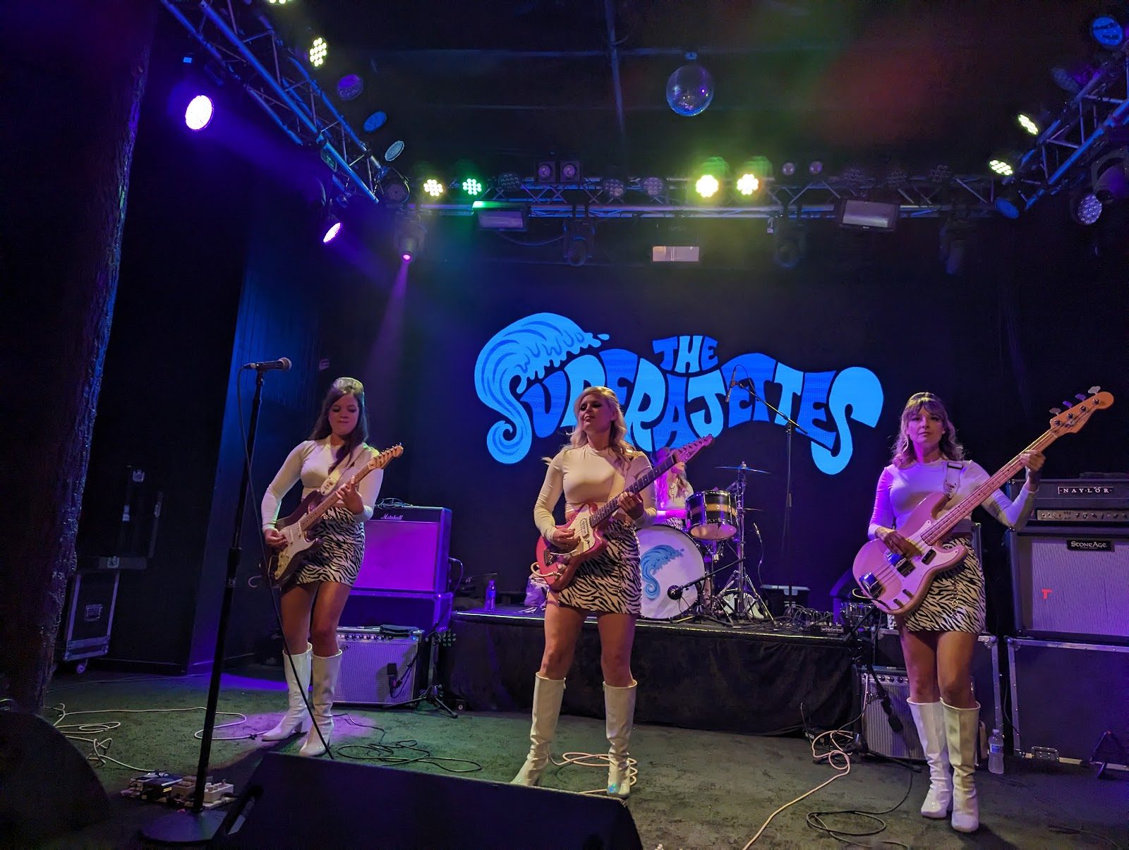 The stage at Trees in Dallas, with a three-piece band in white boots and zebra-print skirts playing under colored lights and a disco ball, the band's logo projected behind them