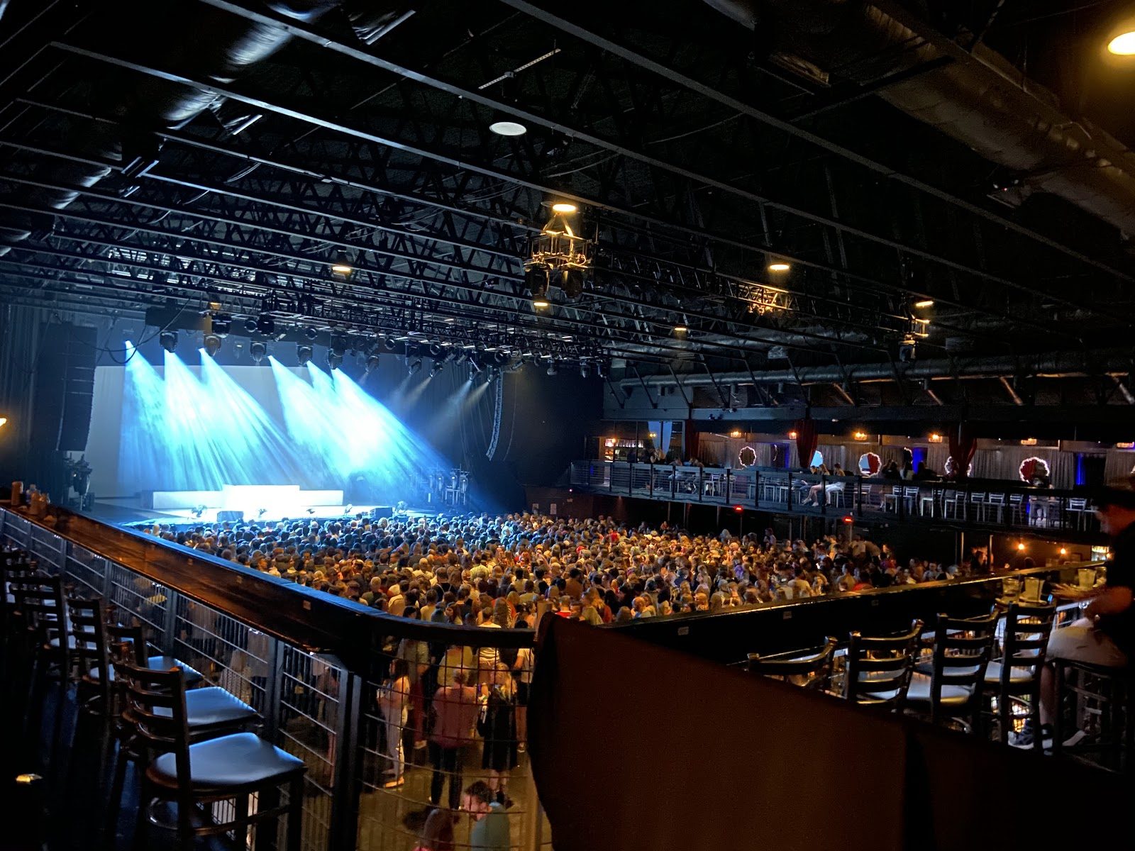 Inside The Bomb Factory in Dallas, seen from the balcony rail: a packed floor crowd facing a stage lit by blue beams under an exposed steel-truss ceiling