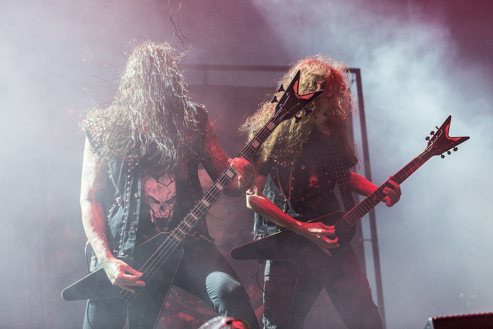 Two long-haired musicians in studded vests playing V-shaped instruments through stage smoke under purple light