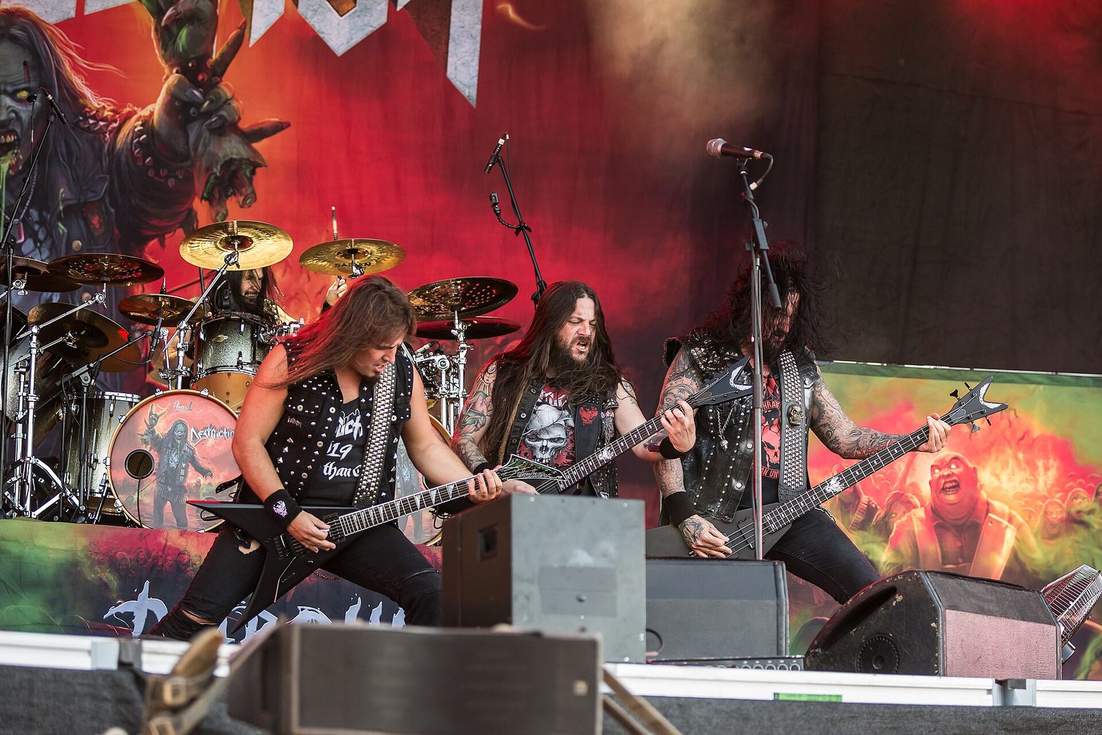 Destruction on an outdoor festival stage: three long-haired players in studded vests with V-shaped guitars and a bass at the front, the drummer behind a kit, a red horror-art backdrop