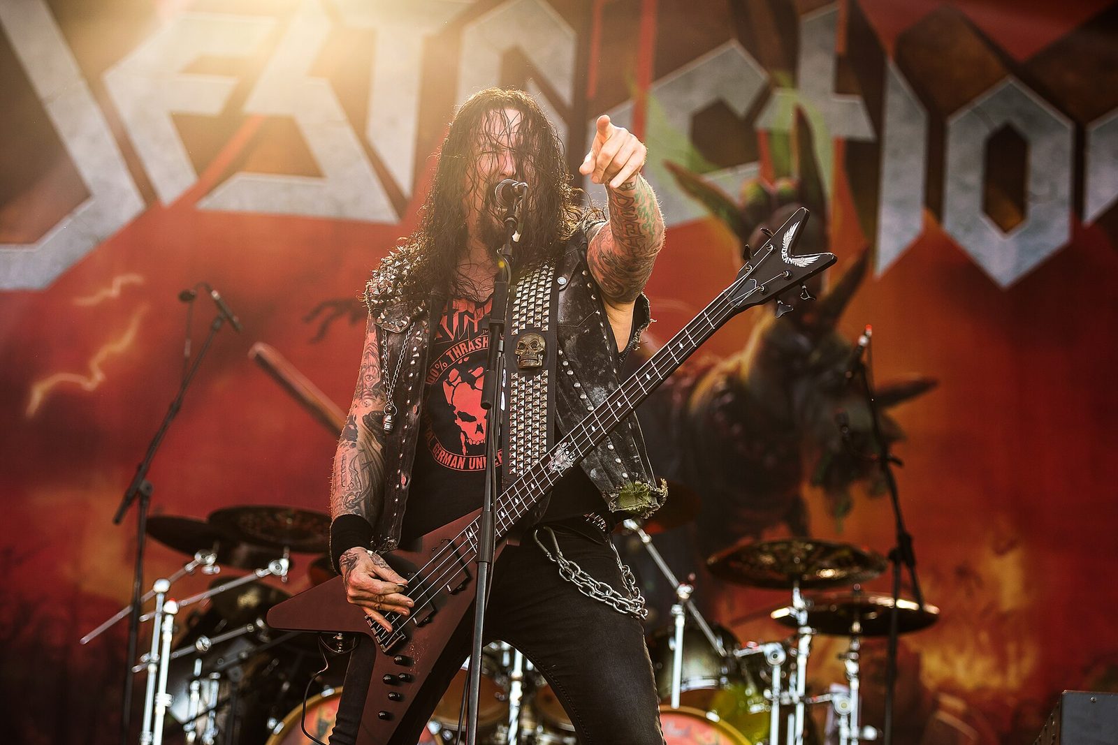 Schmier of Destruction singing into a microphone and pointing at the crowd, long dark hair, a studded vest and a red V-shaped bass, the Destruction logo on the backdrop behind him