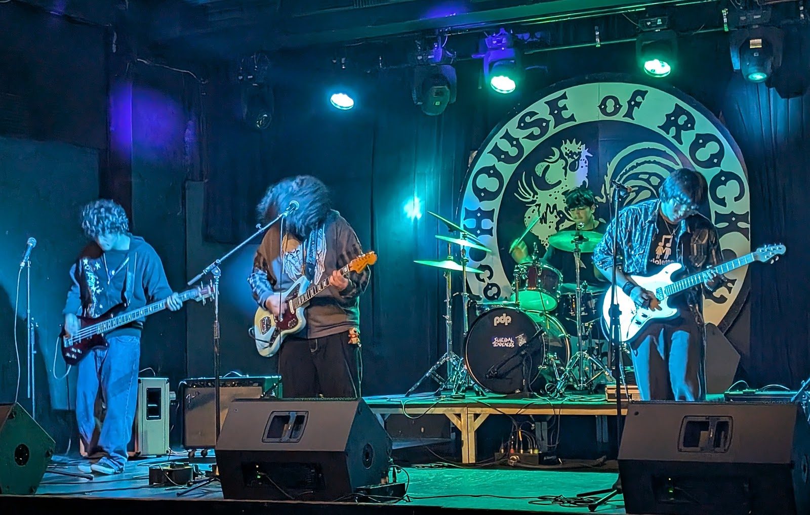 A four-piece band playing under green and blue stage lights in front of the round House of Rock logo backdrop