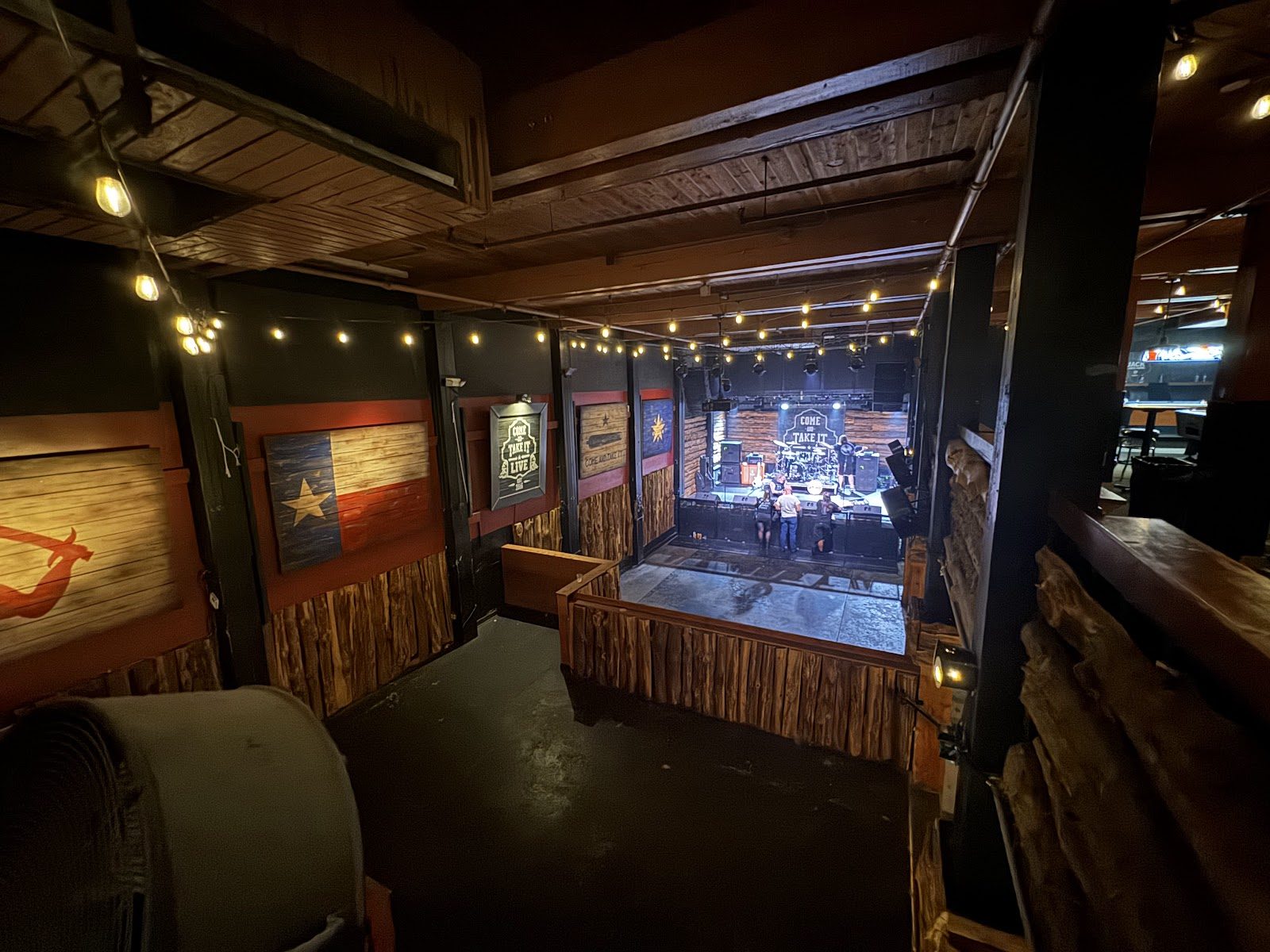 View from a raised wooden walkway down to the stage at Come and Take It Live, with a painted Texas flag on the wall and string lights overhead