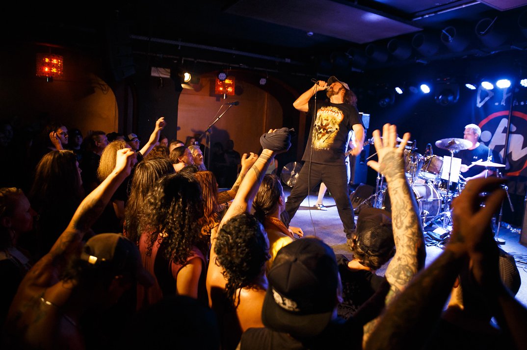 D.R.I. singer Kurt Brecht in a cap and skull T-shirt singing over a club crowd with raised, tattooed arms, the drummer behind him under blue and white stage lights