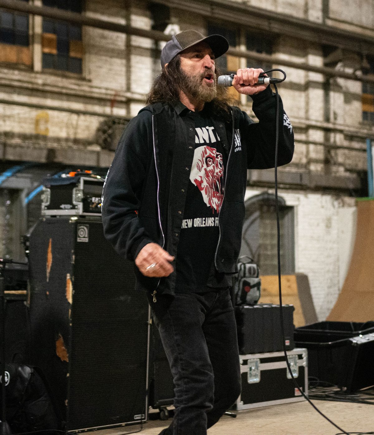 Kurt Brecht, long-haired and bearded in a trucker cap and black hoodie, singing into a handheld microphone in front of amplifiers in a brick warehouse