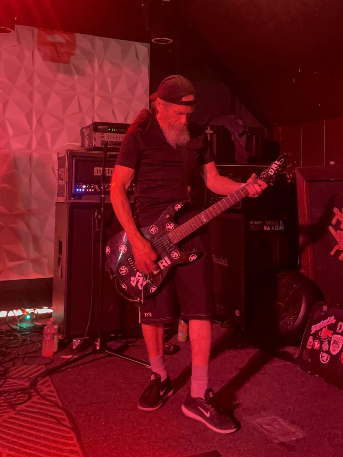 Spike Cassidy, gray-bearded in a backwards cap, playing a black guitar covered in D.R.I. stickers under red stage lights