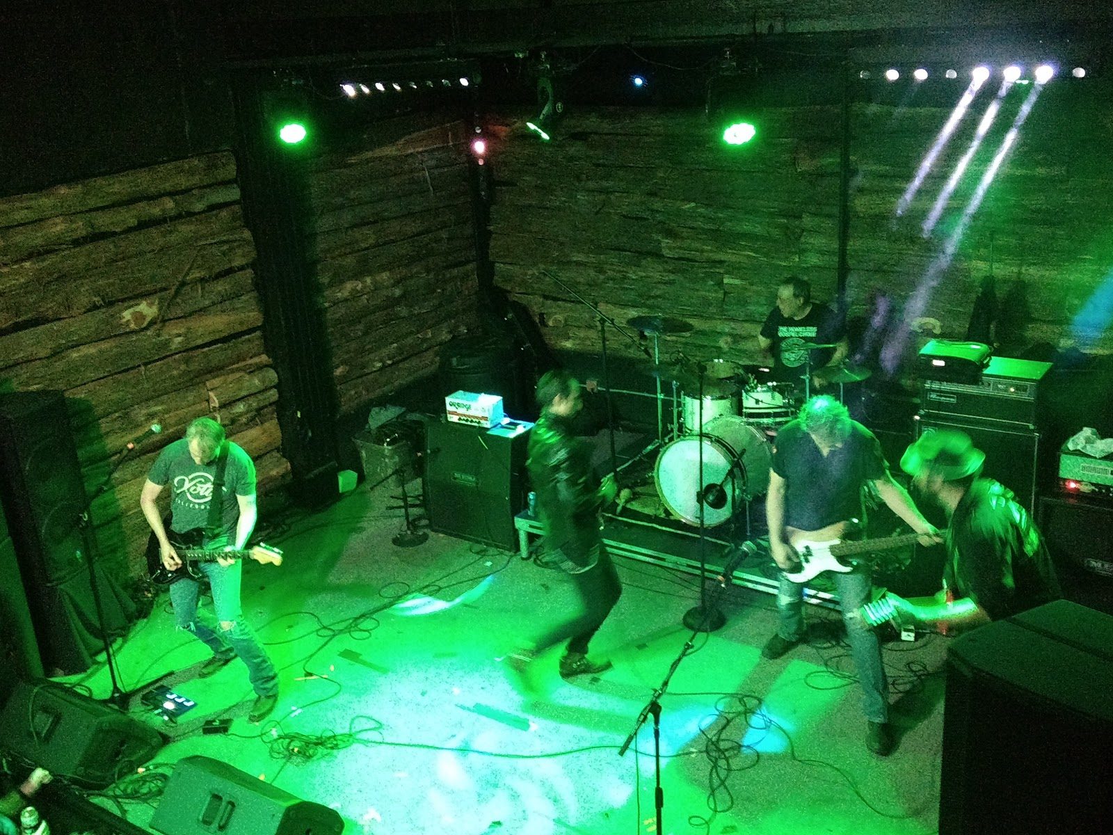 A five-piece band playing under green stage lights at Come and Take It Live, seen from above, against rough wooden plank walls