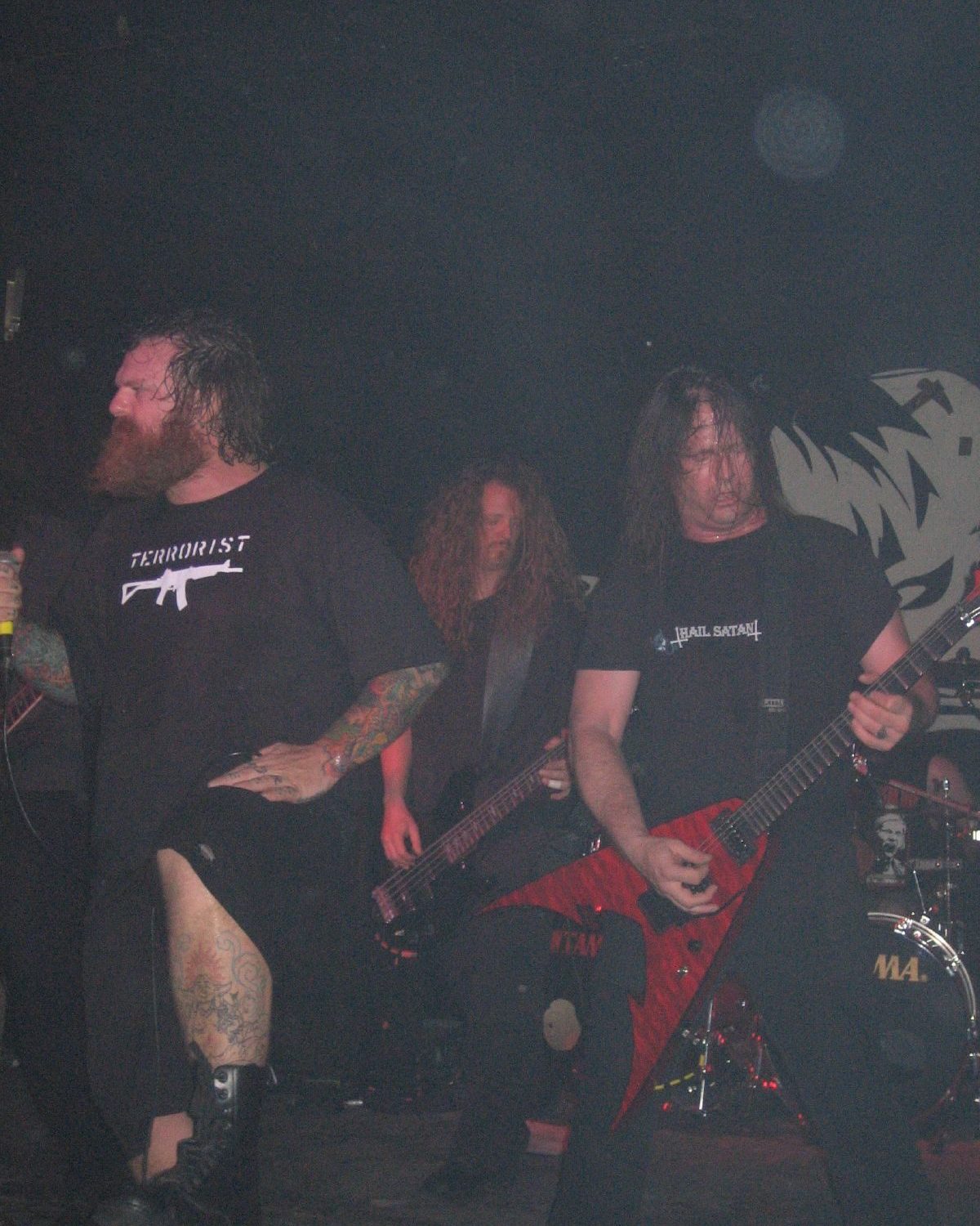 Exodus on a dark club stage: a bearded, tattooed singer at the microphone, a long-haired bassist behind him and a guitarist playing a red V-shaped guitar