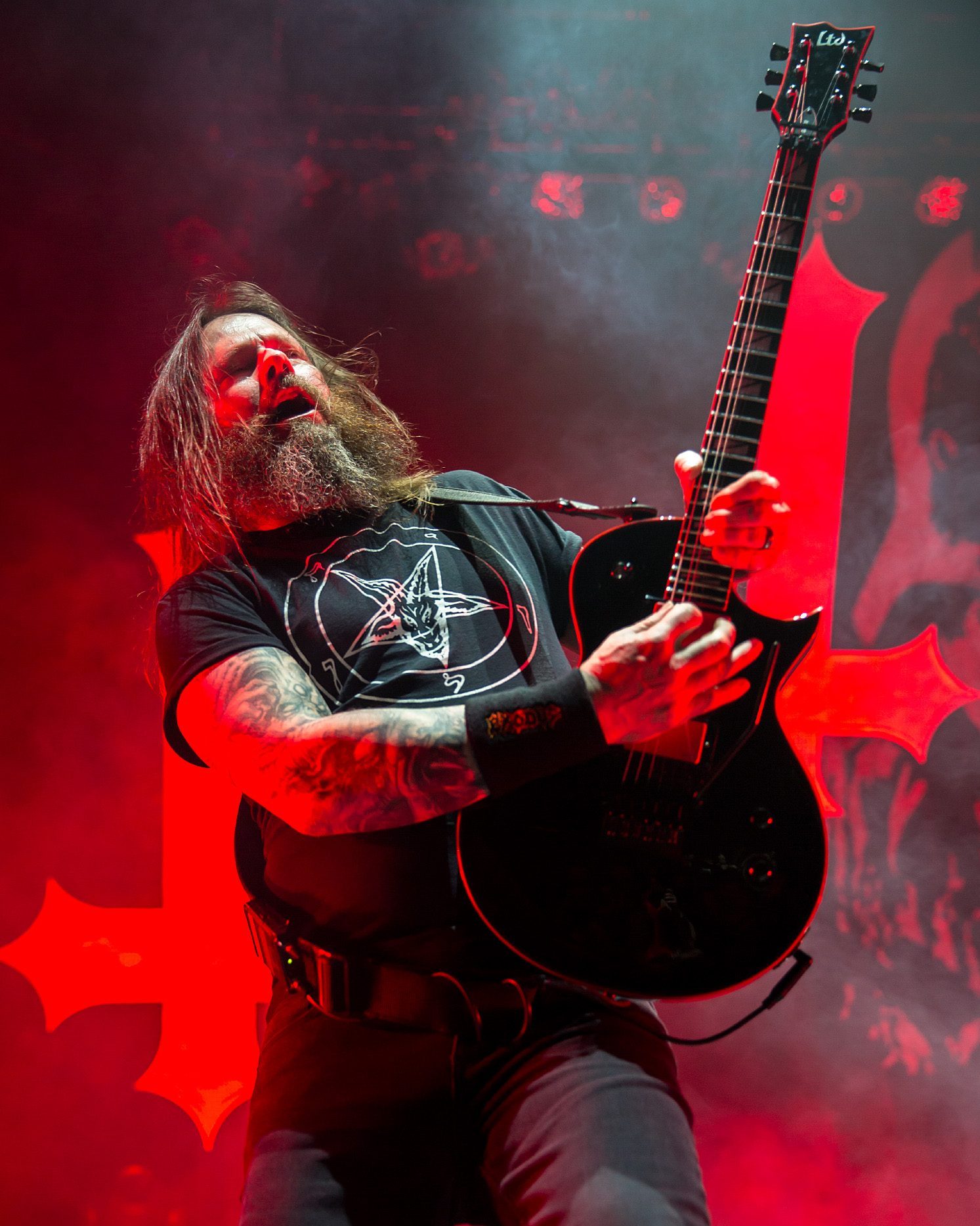 Gary Holt, long-haired and bearded, leaning back with his mouth open while playing a black guitar, a red inverted-cross backdrop behind him
