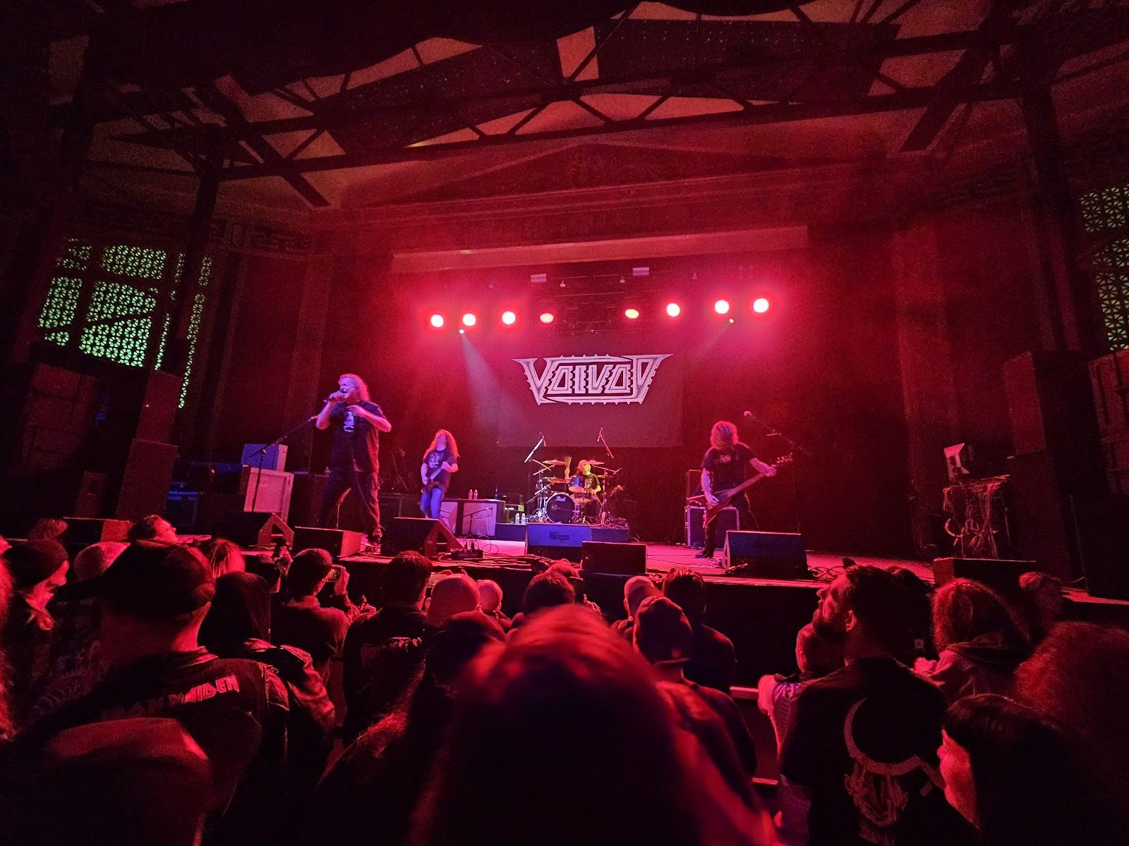 Voivod playing under red stage lights at the UC Theatre in Berkeley, seen from the crowd, with the hall's ornate grillework and ceiling beams above