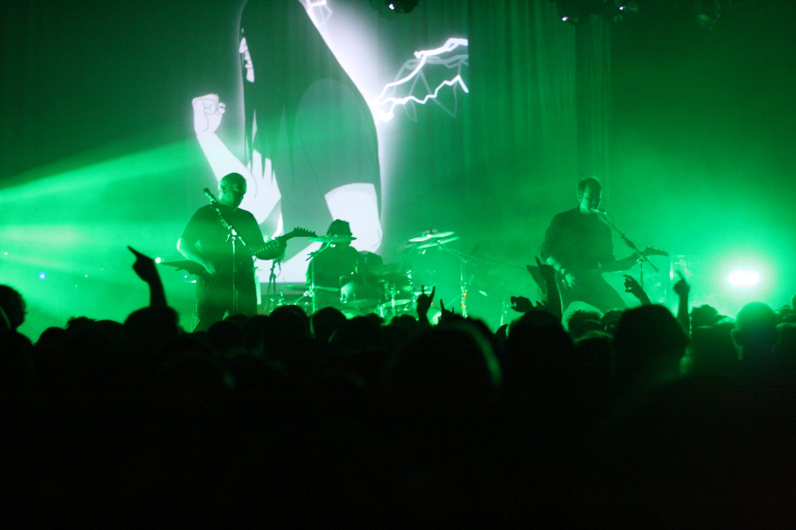 Dethklok on a dark stage lit green, with two guitarists and a drummer in silhouette in front of a large animated projection and a crowd raising their hands
