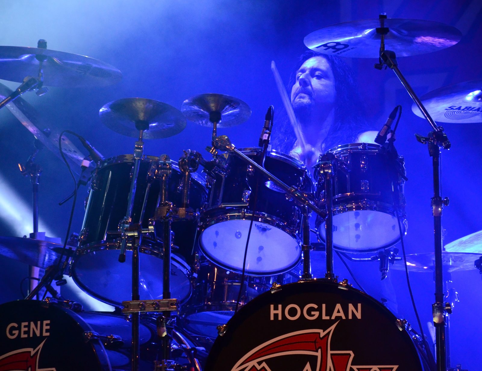 Gene Hoglan raising a drumstick behind a chrome drum kit under blue light, with a bass drum head reading HOGLAN in front