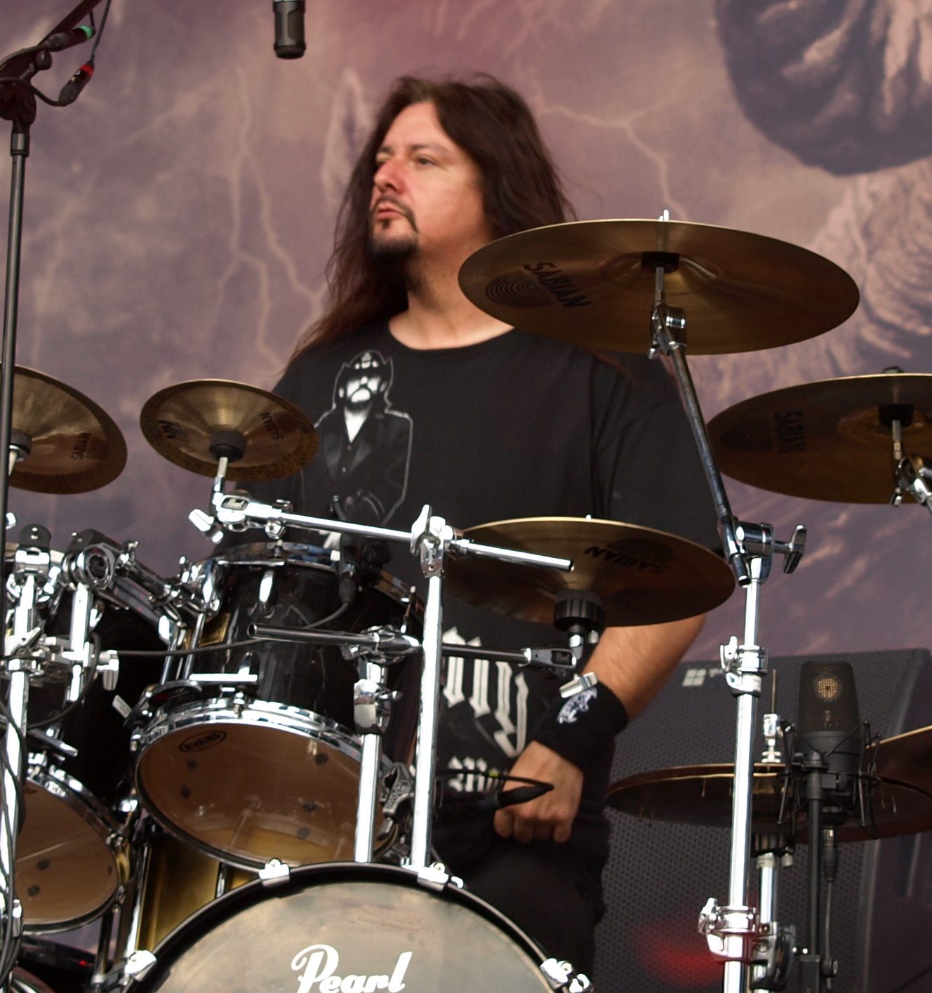 Gene Hoglan in a black T-shirt behind a kit with Sabian cymbals and a bass drum head reading Pearl, in front of a painted backdrop