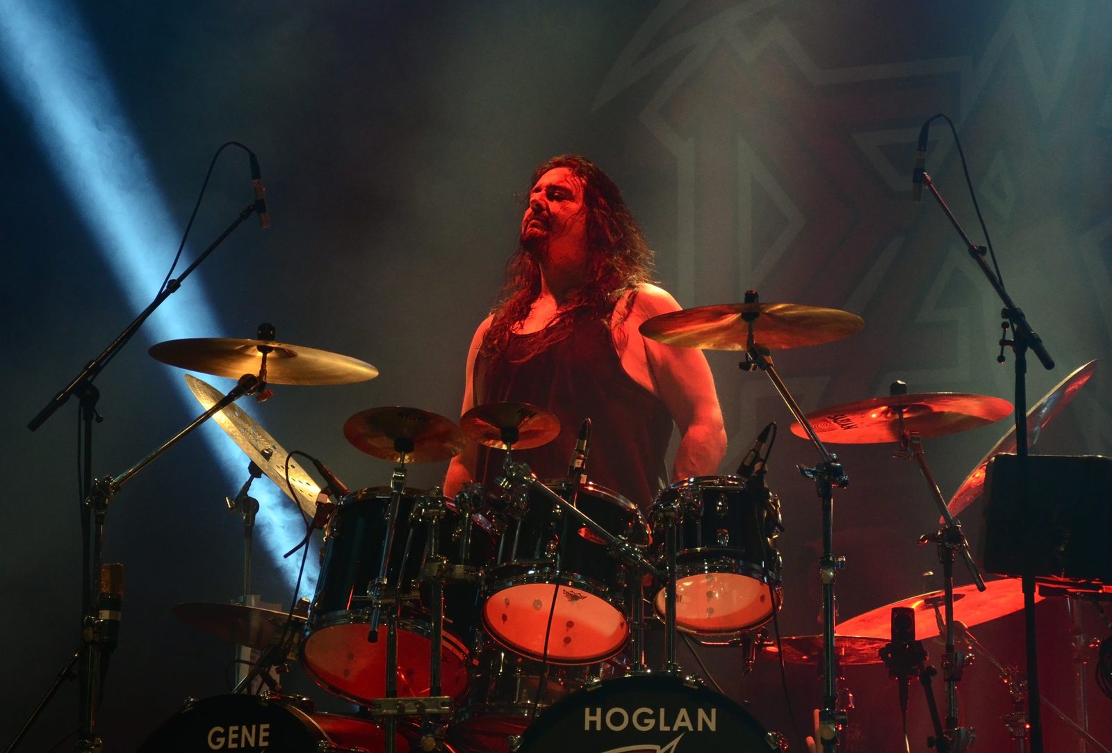 Gene Hoglan, with long dark hair and a black tank top, head tilted back with eyes closed behind a black drum kit with bass drum heads reading GENE and HOGLAN, lit red with a blue beam crossing the stage