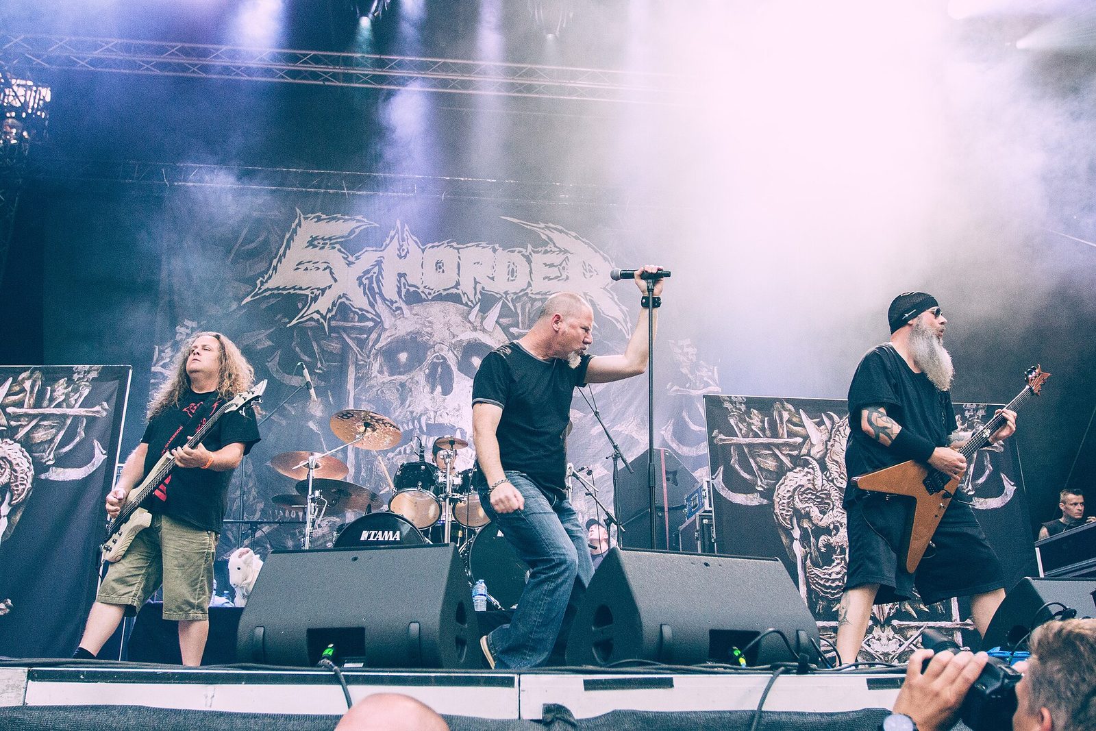 Exhorder on an outdoor festival stage: a bassist with long curly hair, a singer in a black T-shirt holding the mic stand overhead, and a bearded guitarist with a V-shaped guitar, in front of a giant Exhorder skull backdrop