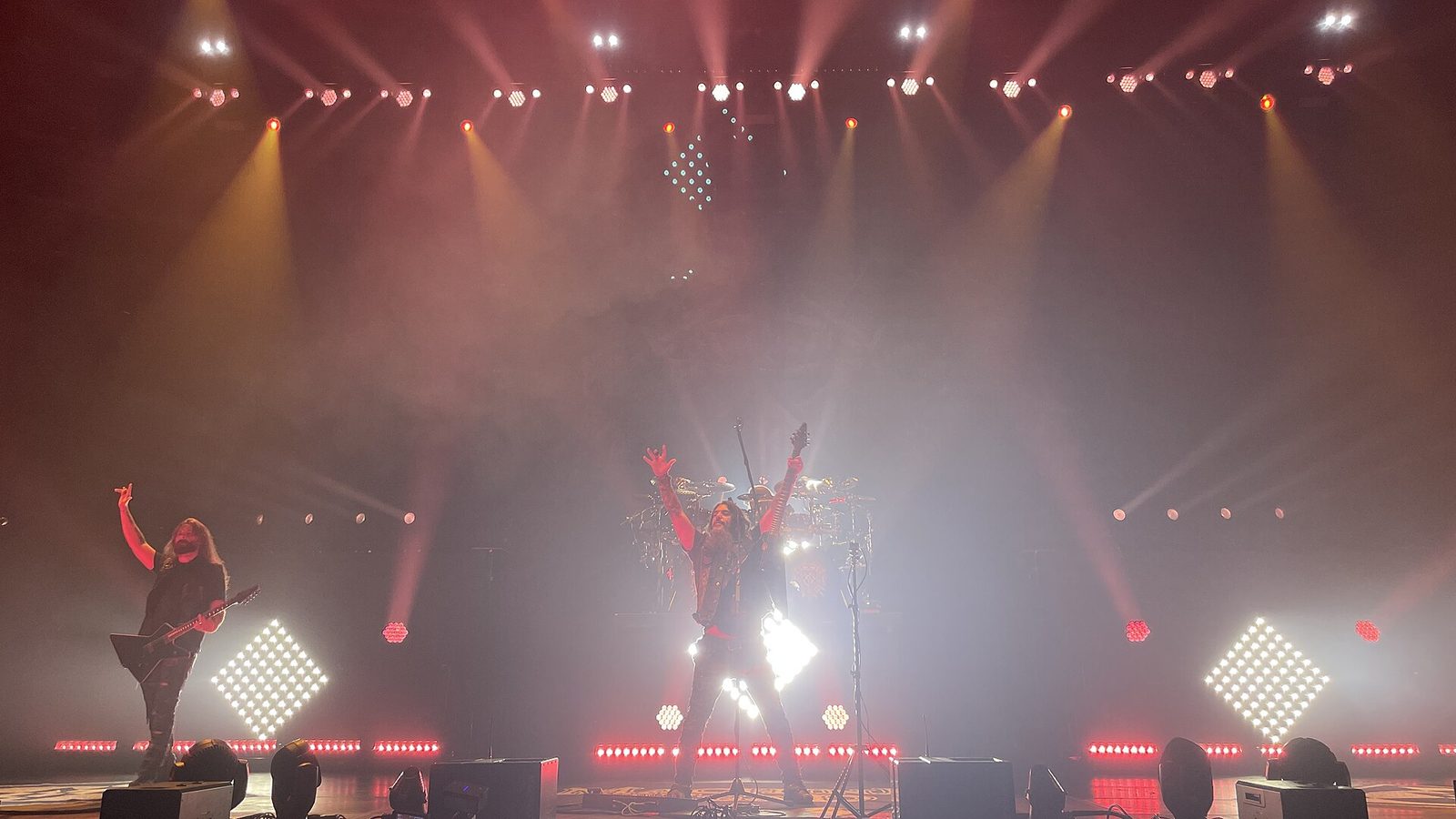 Two members of Machine Head raising their arms on a smoky stage under red and white lights, with a drum kit behind them