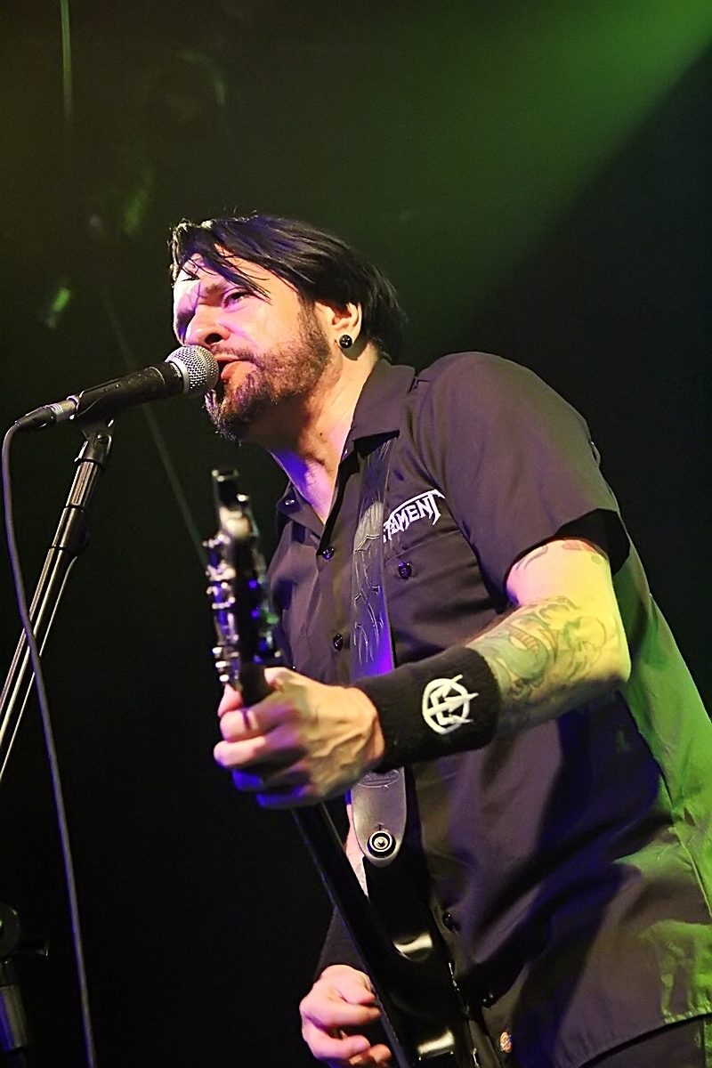 Tommy Victor, with dark hair and a beard, singing into a microphone while playing guitar under green stage light