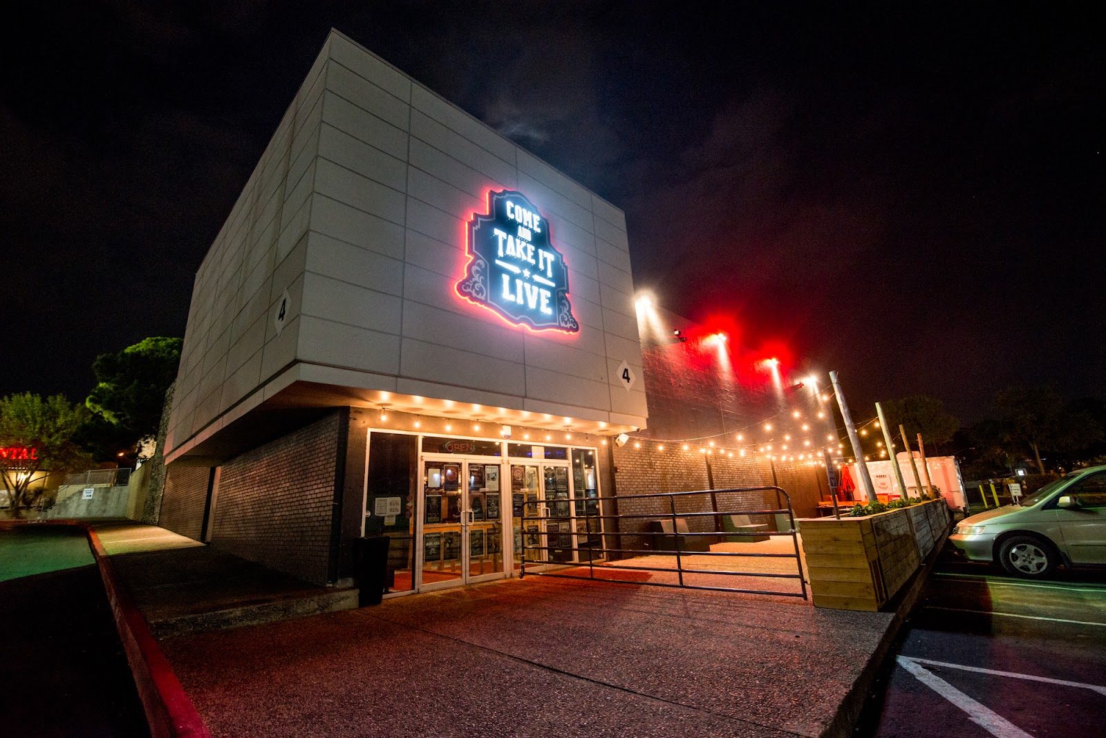 Come and Take It Live at night, its red-lit sign on a gray building marked 4 above glass doors, with string lights over the patio