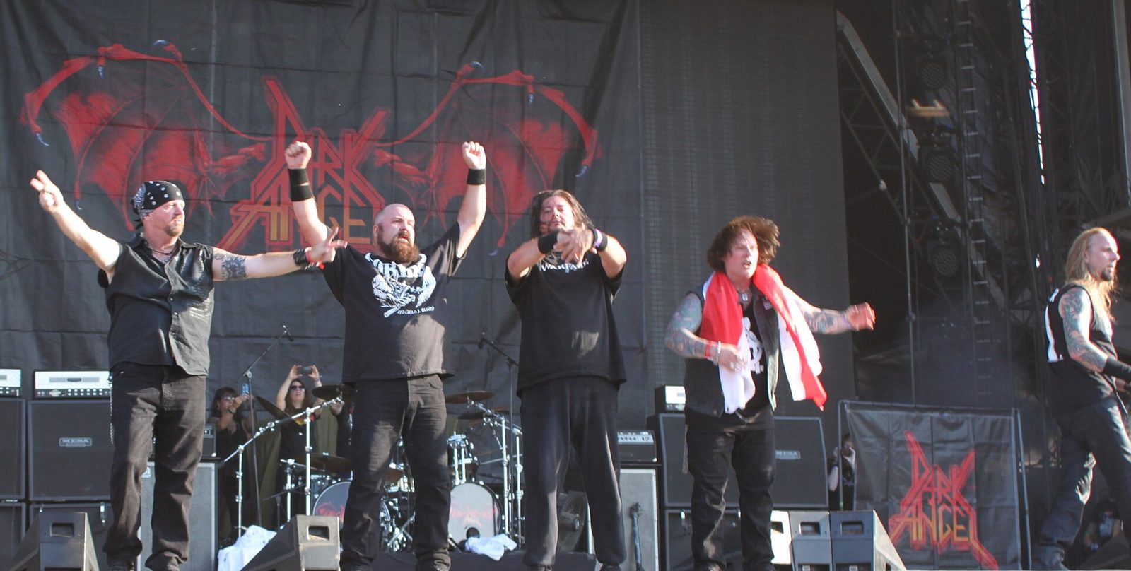 Five members of Dark Angel stand at the front of a large outdoor festival stage with arms raised, under a red Dark Angel banner