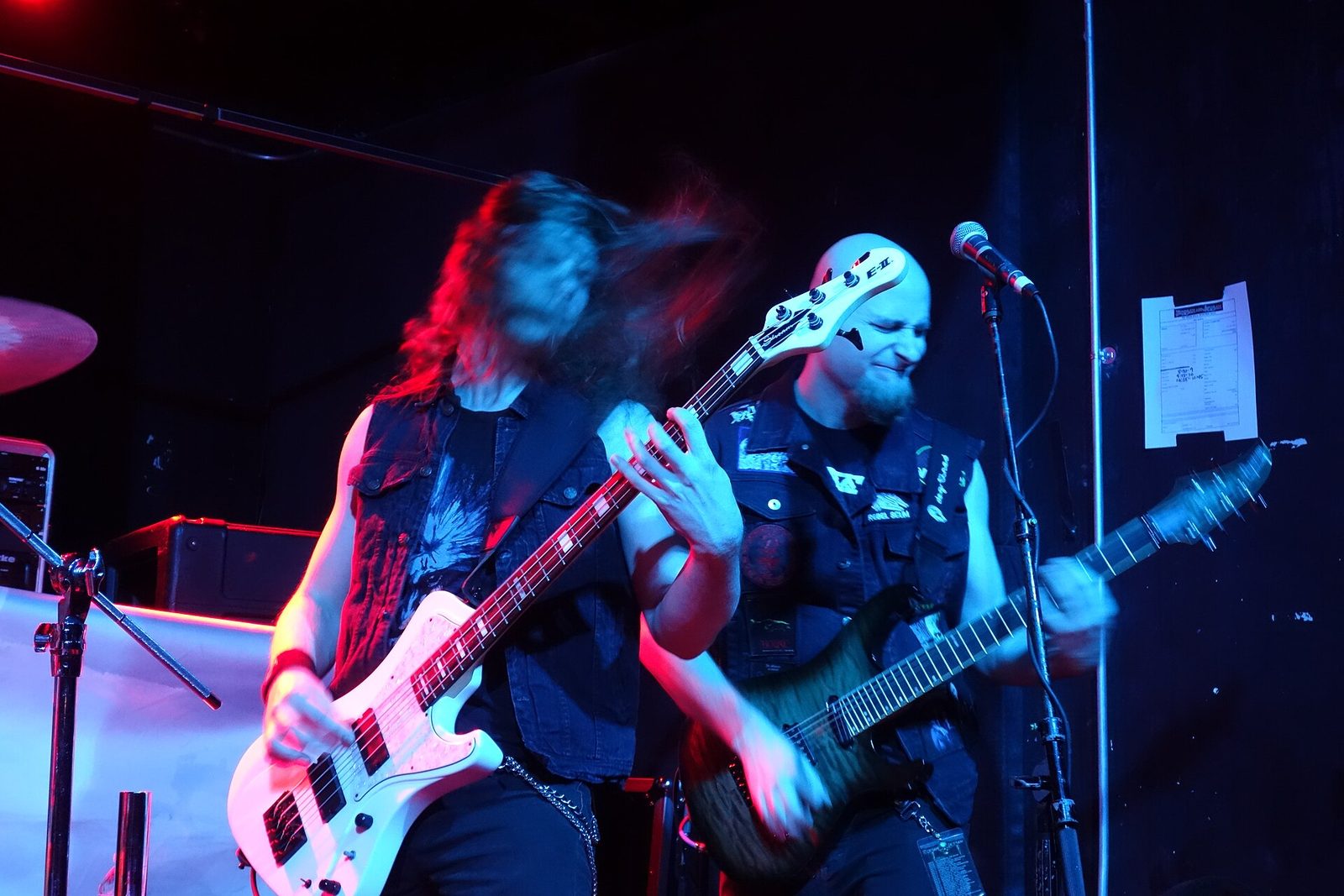 Helstar's bassist with a white bass and guitarist with a dark guitar play side by side under blue and red club lights, the bassist's long hair flying