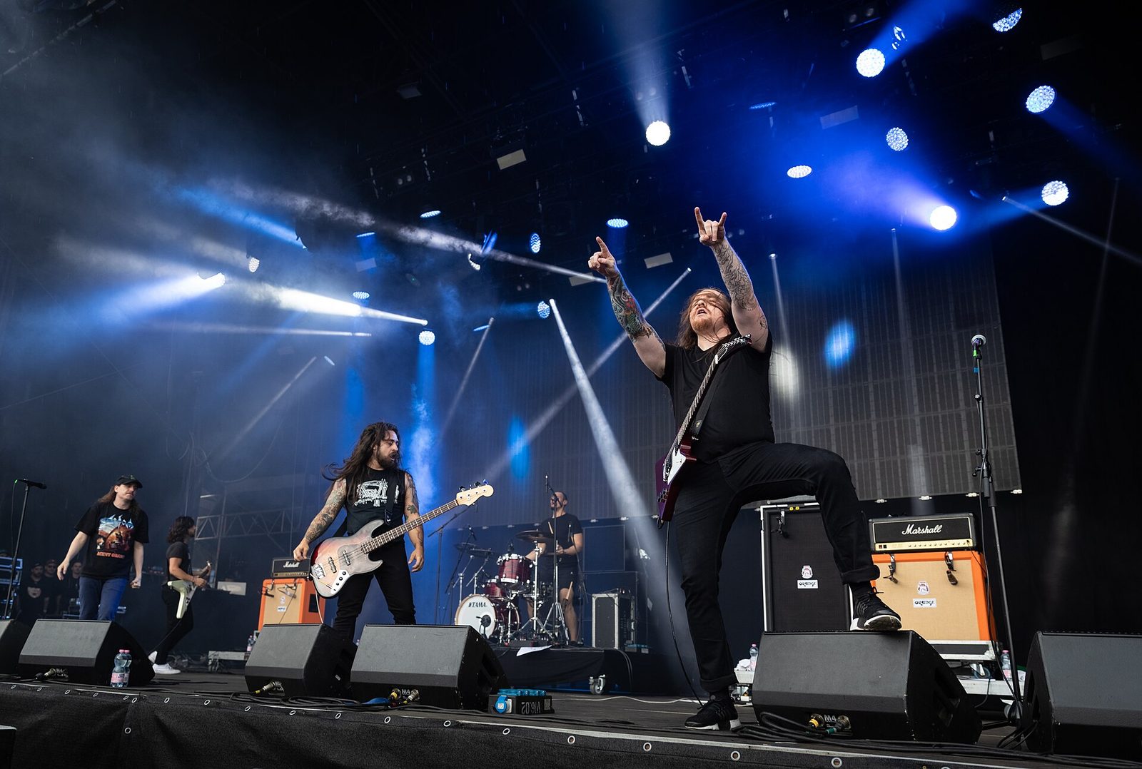 Power Trip on a large outdoor stage under blue lights, a guitarist with one foot on a monitor raising devil horns while the bassist and drummer play behind him