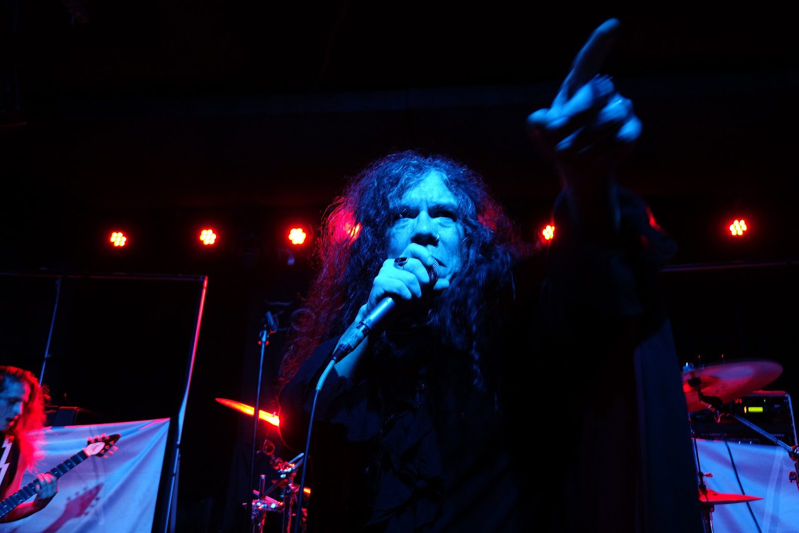 James Rivera singing under blue and red stage light, curly hair to his shoulders, pointing out at the crowd while a guitarist plays at the left edge