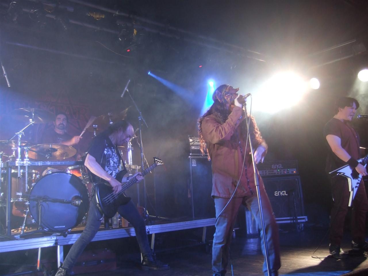 Helstar onstage in Germany in 2009: a masked singer with long hair at the microphone in smoky white light, a bassist leaning into his playing, a guitarist with a white flying-V at right and the drummer behind