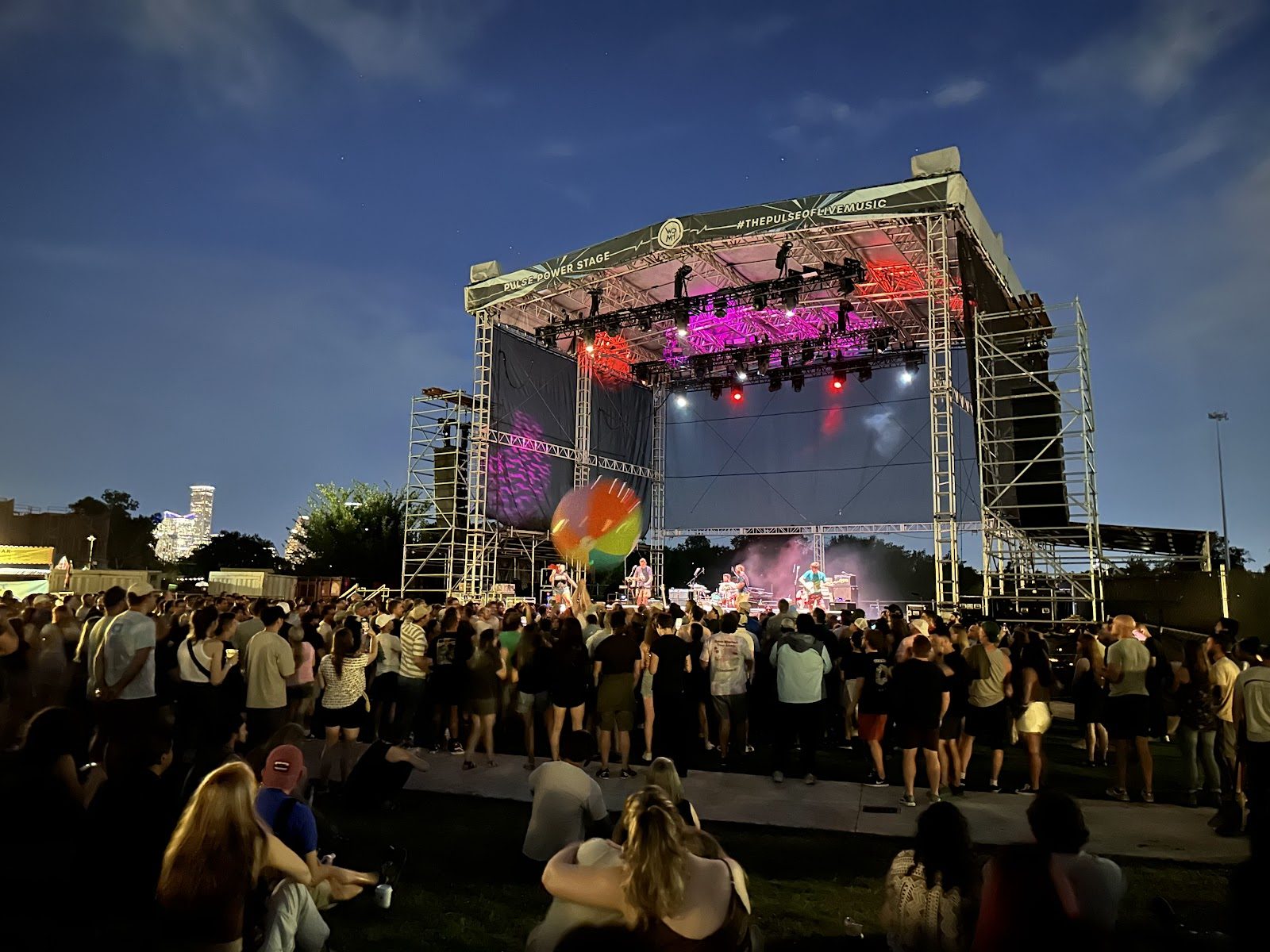 Crowd on the lawn at White Oak Music Hall at dusk facing a big outdoor stage with pink and red lights, and the downtown Houston skyline to the left