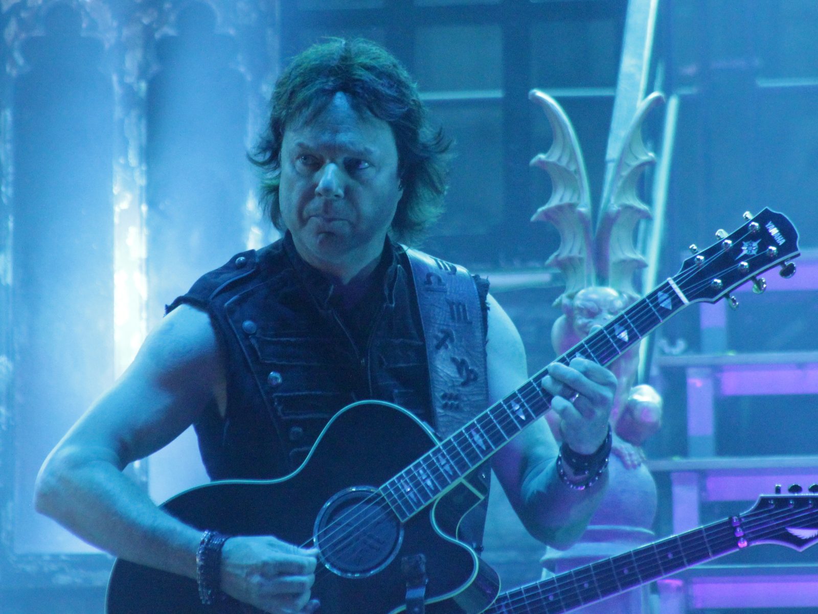 Andy LaRocque, with shoulder-length brown hair and a sleeveless black top, playing a black acoustic guitar on a blue-lit stage with gothic set pieces behind him
