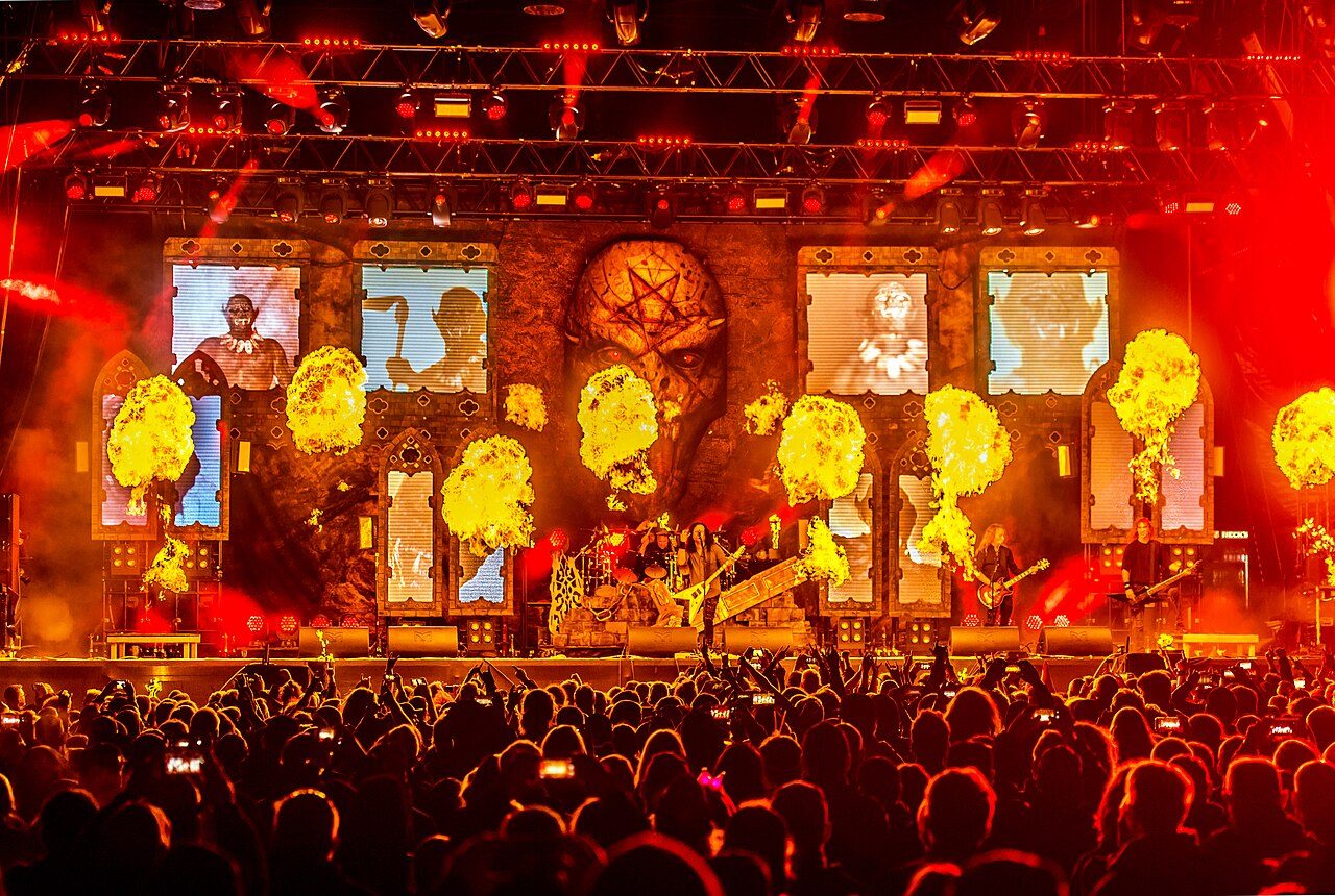 Kreator on a huge festival stage at night, flame bursts rising in front of a backdrop of painted demon panels, with a packed crowd silhouetted in the foreground
