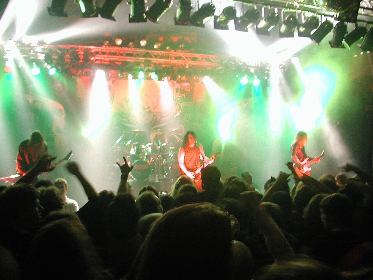 Kreator on a club stage washed in green and white light, three long-haired musicians with guitars and a drum kit in the middle, fans' raised hands in the foreground
