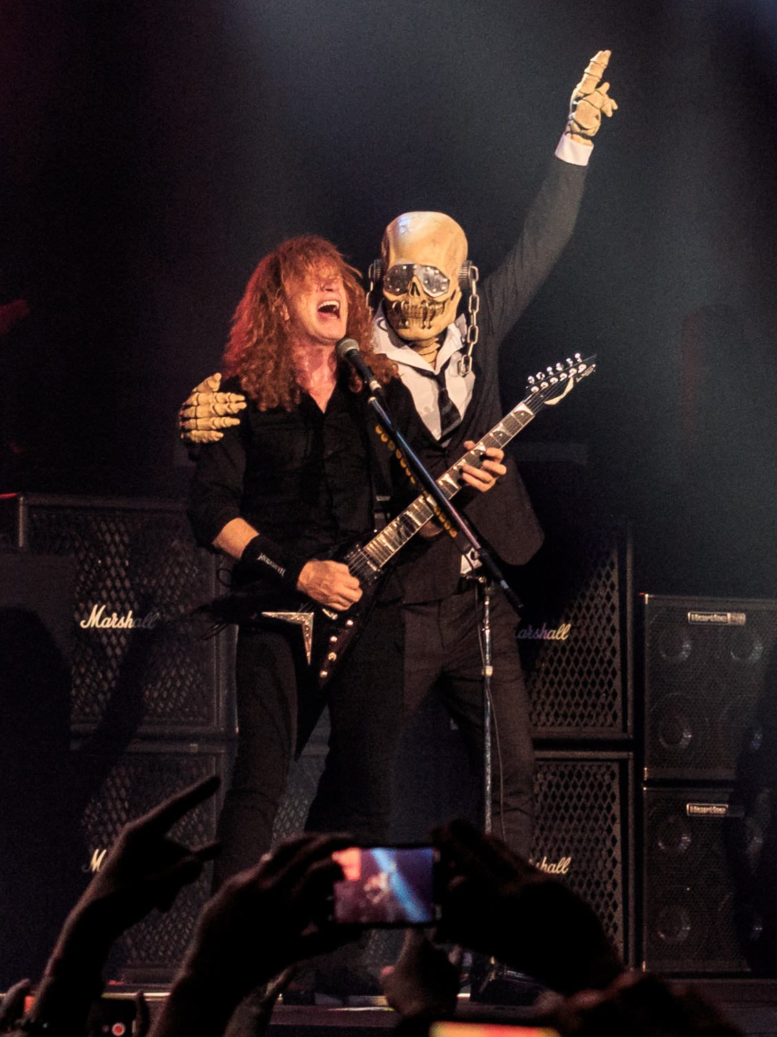 Dave Mustaine playing a black flying-V guitar while a costumed Vic Rattlehead in a black suit and skull mask stands behind him with a hand raised