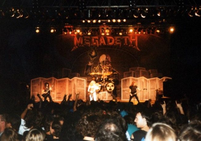 A darkened arena stage lit orange, with a large Megadeth logo and a giant skull figure above the drum riser, four musicians on stage and raised hands in the crowd
