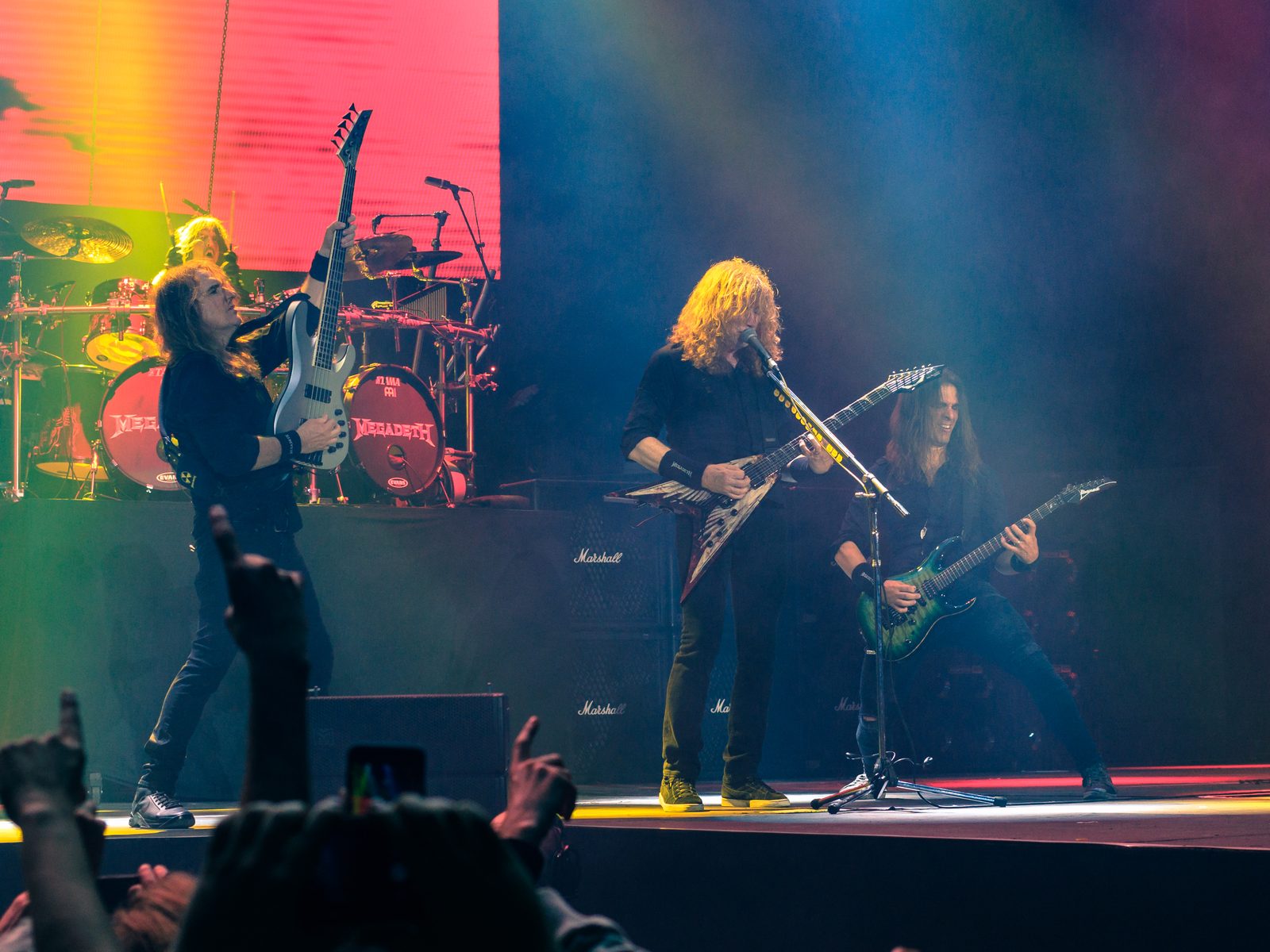 Megadeth on a red and blue lit arena stage, Dave Mustaine at center with a flying-V guitar, a bassist to his right and a guitarist with a green guitar to his left, drums and Marshall stacks behind