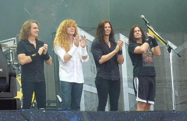 Four members of Megadeth standing in a row at the front of an outdoor festival stage, applauding the crowd, Dave Mustaine second from left in a white shirt