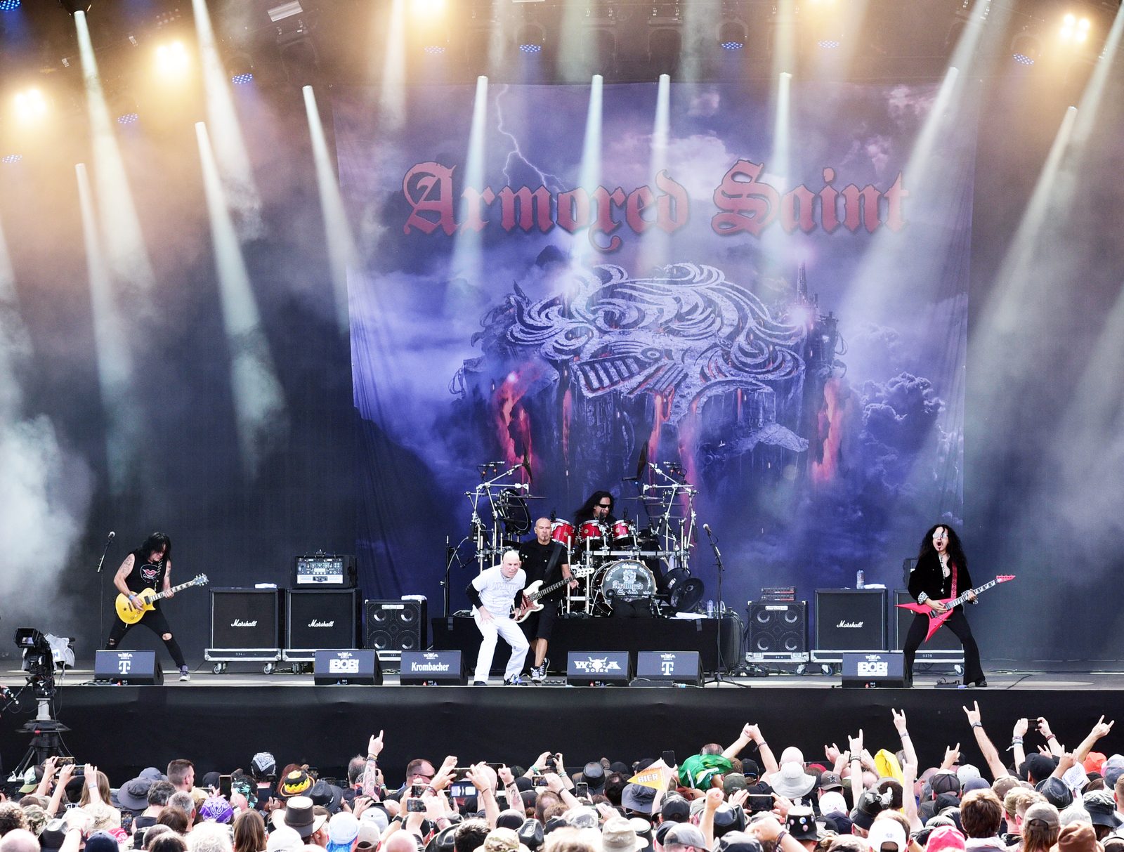 Armored Saint performing on a festival stage under white spotlights in front of a large backdrop with the band's gothic logo, a crowd raising devil horns in the foreground