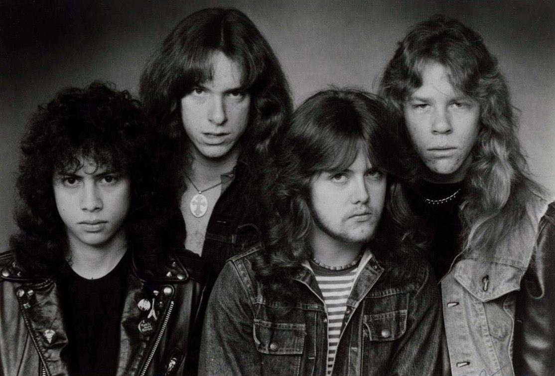 Black-and-white 1983 press photo of Metallica's four members standing close together in leather and denim jackets, all with long hair, staring at the camera